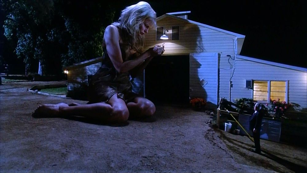 A scene from Attack of the 50ft Woman (1993). Nancy, now a giant, holds and tries to read a normal-sized contract under a street light.