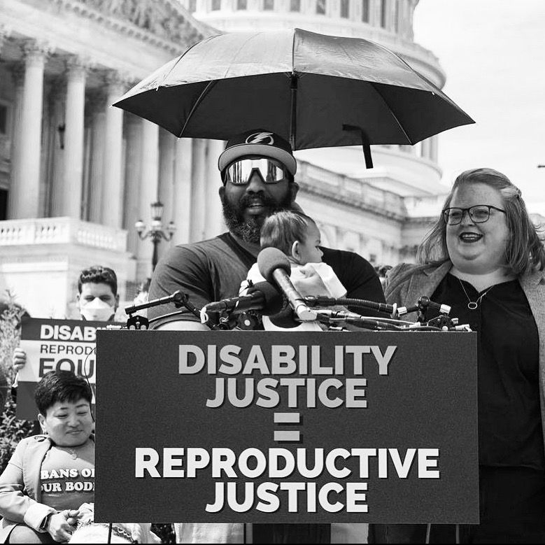 Black and white photograph of AJ standing at a press lectern outside the Capitol holding a baby. The lectern has a sign that says “Disability Justice = Reproductive Justice.” Tory is next to him holding an umbrella above his head.