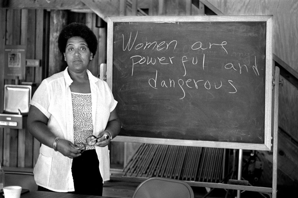 Black and white photograph of Audre Lorde standing in front of a chalkboard with the handwritten words “Women are powerful and dangerous” on it.