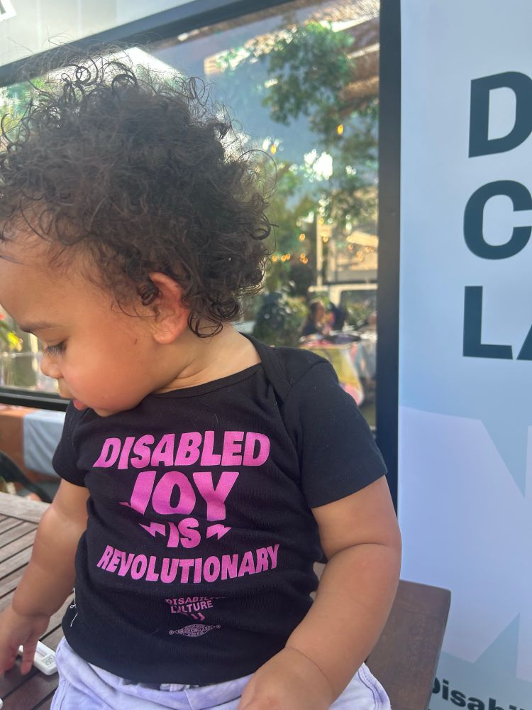 Baby Kink wearing a black shirt with magenta text that says Disabled Joy is Revolutionary. 