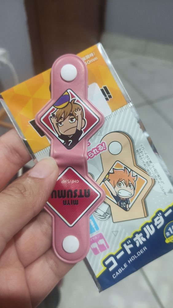 Foto de uma mão branca segurando a embalagem e um organizador de cabos do anime Haikyuu. O organizador tem a imagem do personagem Miya Atsumu.
