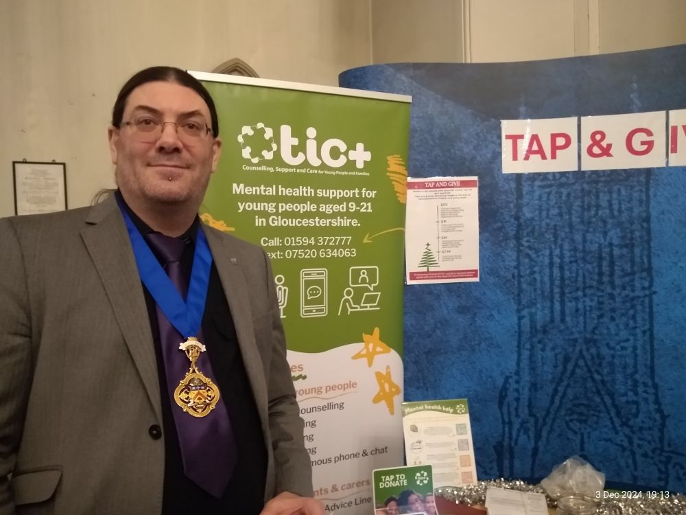 Cllr David Willingham, wearing the Deputy Mayor of Cheltenham's badge of office standing next to a banner about the work of TIC+ http://www.ticplus.org.uk/