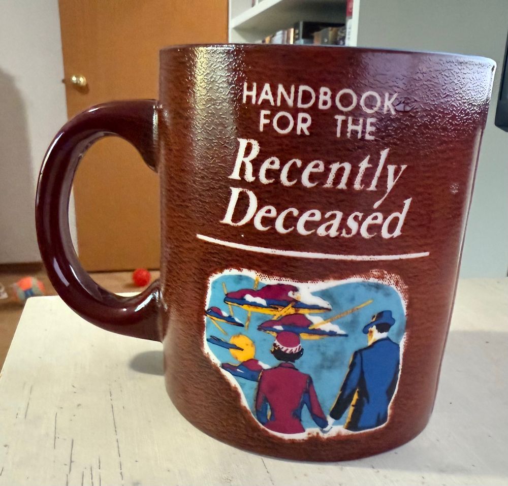 A brown mug with a reference to the movie Beetlejuice that says “handbook for the recent deceased.” A man and woman are holding hands and walking into the light below the wording
