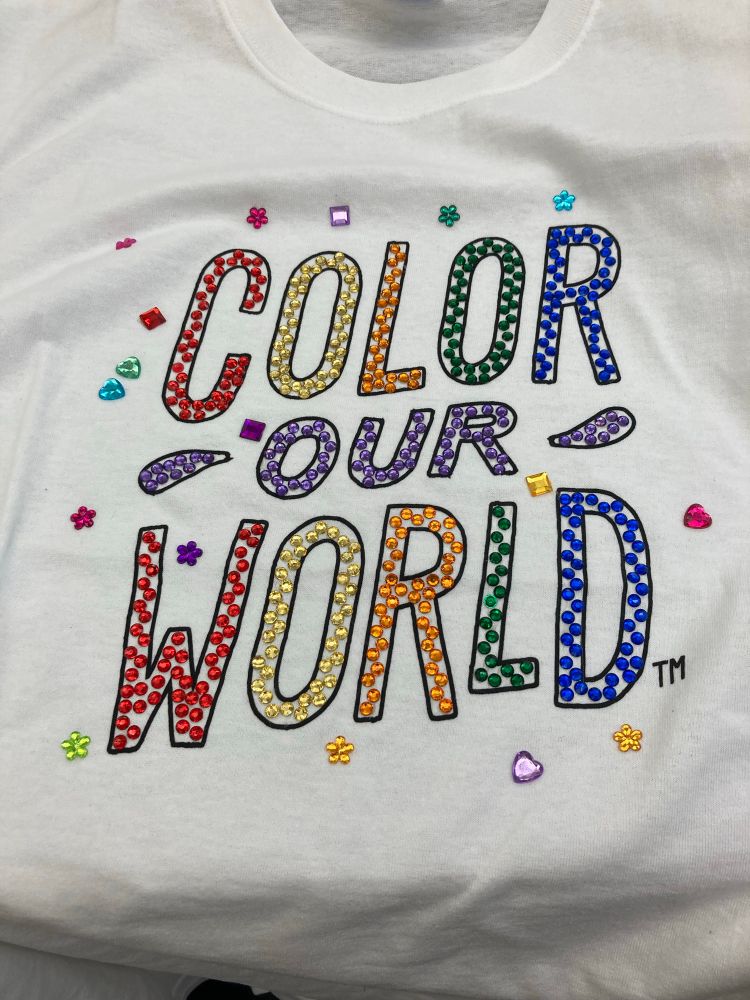 Bedazzled letters spelling out color our world in a rainbow pattern with heart and flower accents 