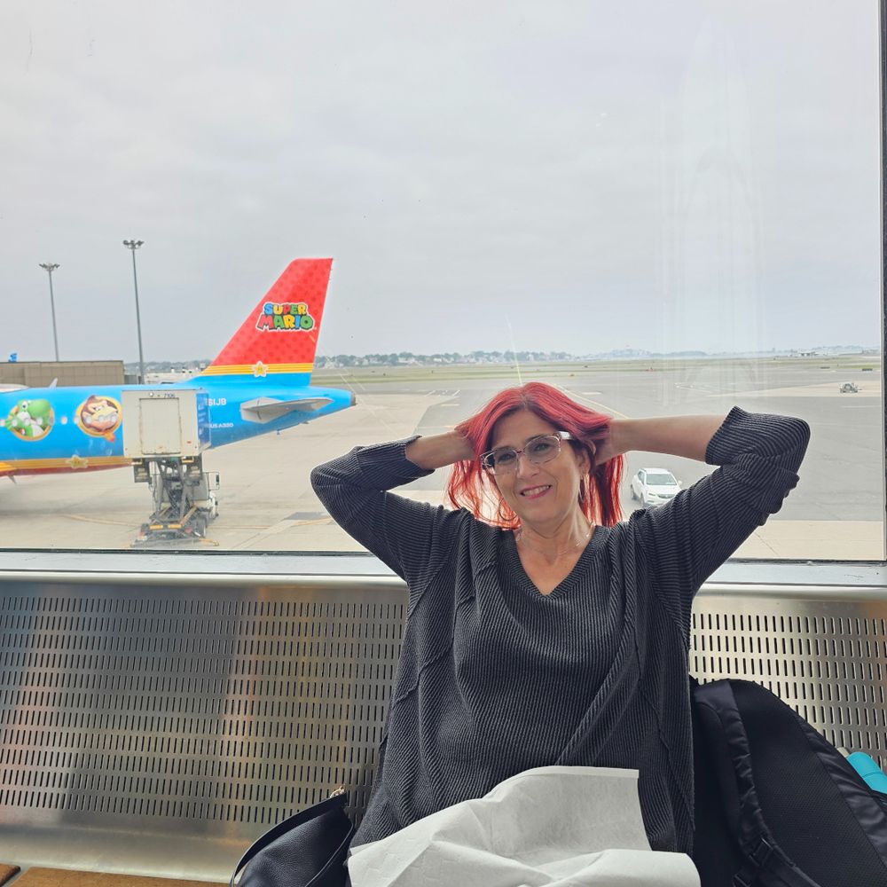 Stacey leaning back on a metal bench at an airport. An airliner pained for Super Mario can be seen out the window.