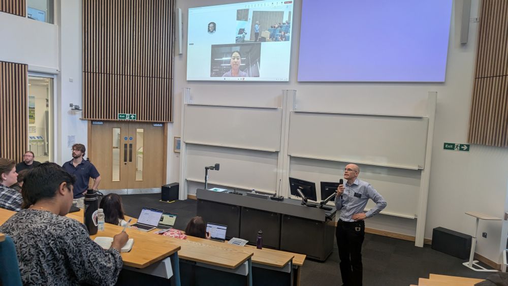 Mandy Xiang speaks remotely to a crowded lecture theatre at Lancaster University at the Device Prototyping Summer School. Professor Steve Hodges takes questions at the front of the room while Mandy's face is displayed on a large screen.