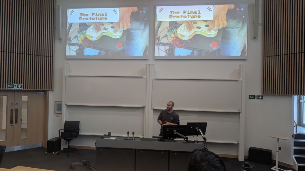Jack Burnett stands at the front of a lecture theatre with a desk before him and two large screens showing a PowerPoint slide above him. The slide shows a prototype of a video game controller and the text 'The Final Prototype'.