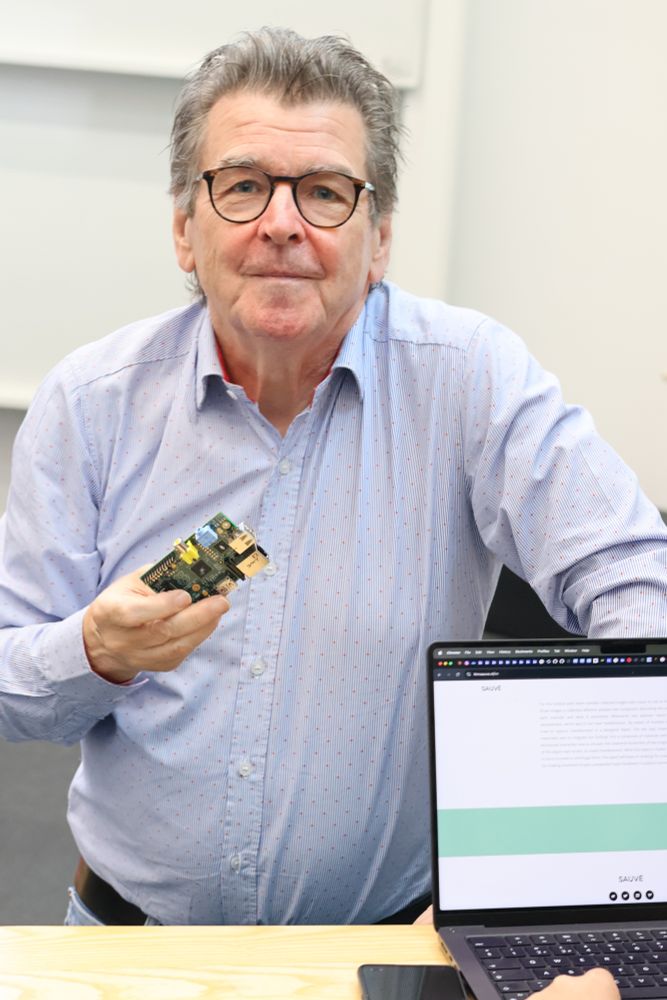 Pete Lomas stands in front of a desk in a lecture theatre with a laptop before him, holding a Raspberry Pi and looking toward the camera.