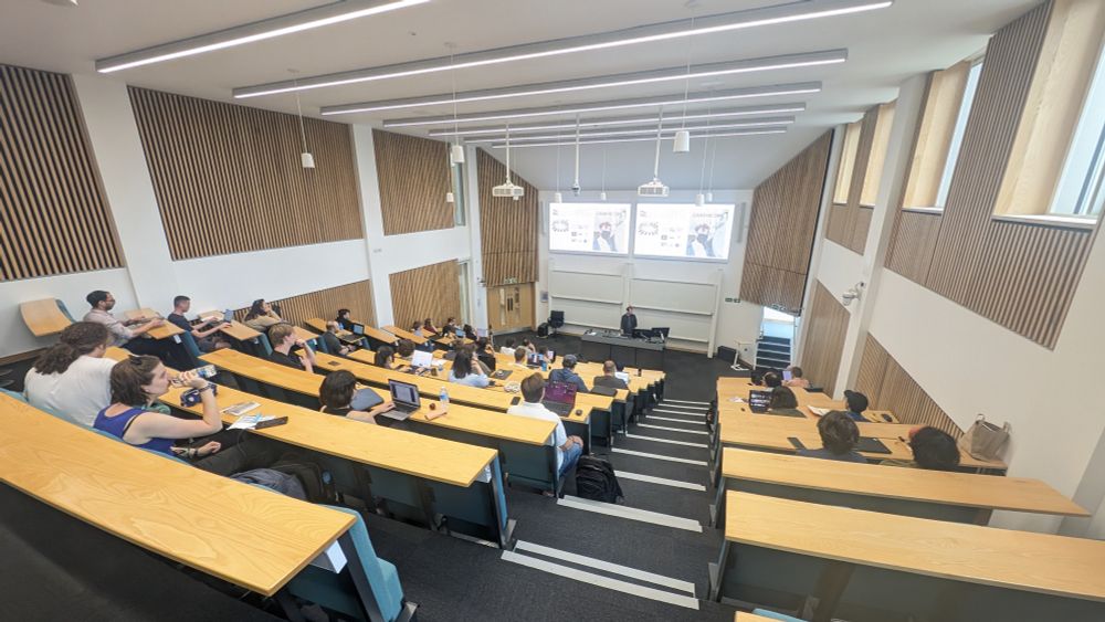 A photo shows a crowded lecture theatre with wood panelled walls at Lancaster University. A presenter stands at a desk at the front of the room with two large display screens overhead showing PowerPoint slides.