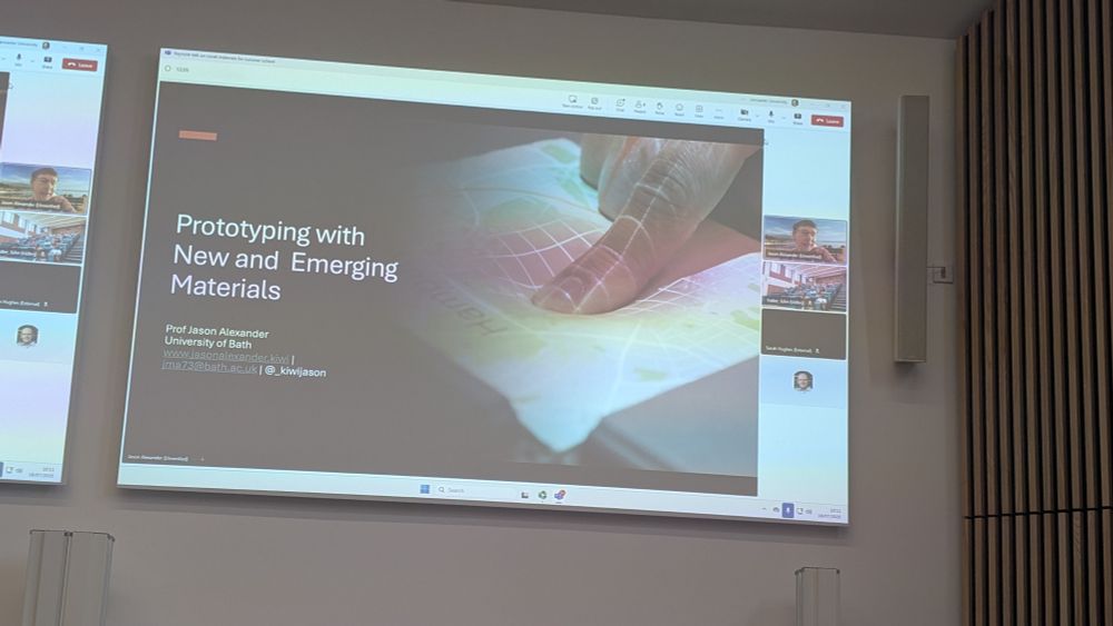 Photo of a large screen displaying a PowerPoint slide, with remote video conferencing displaying Professor Jason Alexander, a classroom, and Steve Hodges to the right. The text on the slide reads 'Prototyping with New and Emerging Materials'.