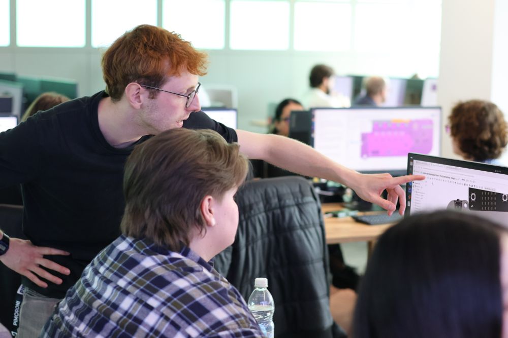 Oliver Child, wearing a black T-shirt and glasses, stands next to a summer school delegate in a computer lab, pointing at something on a computer screen during a practical tutorial.