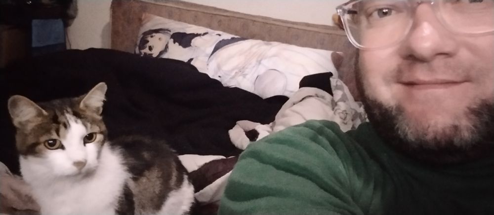 Cat sitting on bed behind a guy with a beard and glasses, smiling