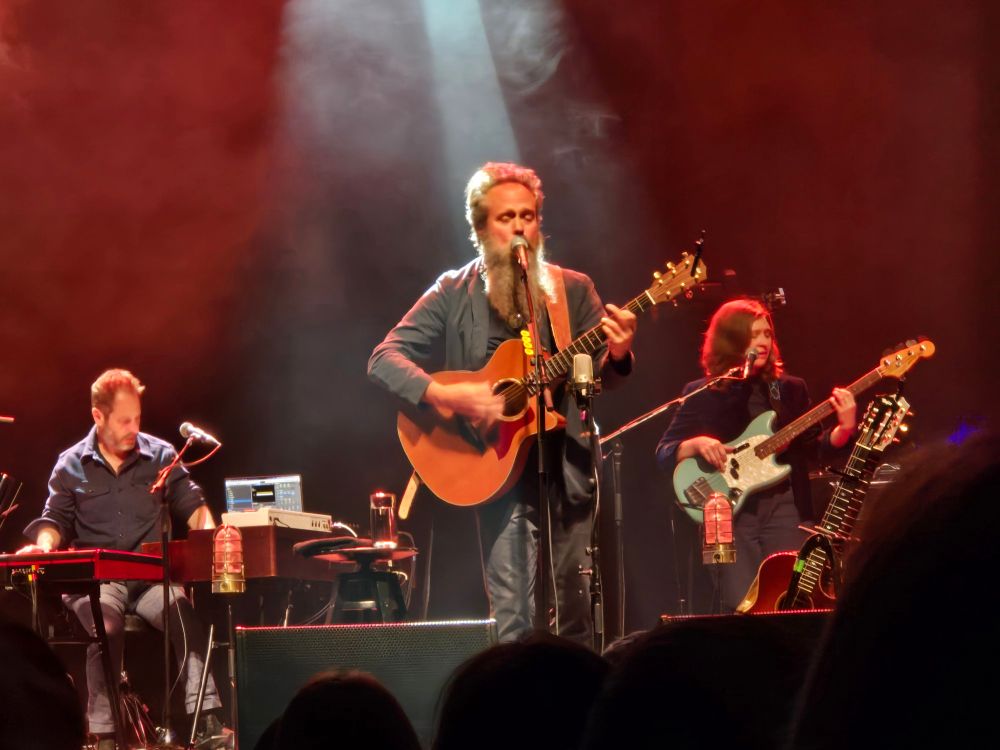 a picture of sam beam from the band "iron & wine" singing at a microphone while playing an acoustic guitar. two bandmates behind him play on an electric guitar while the other is at the keyboards