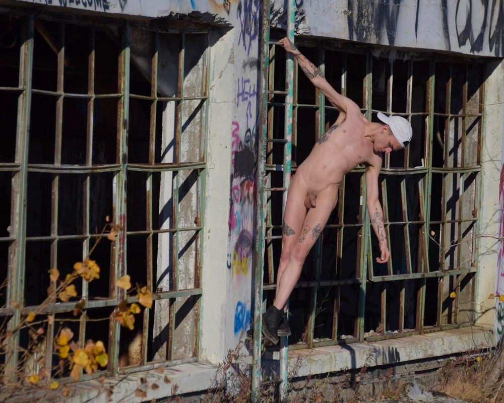 Naked male model hanging off of a ladder in front of broken windows