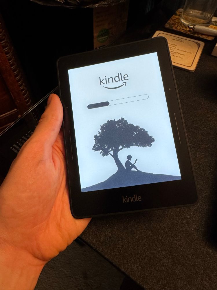 Jack holds an Amazon Kindle Voyage (2014). It displays a loading screen with the Kindle logo above a progress bar, below which is a black-and-white illustration of a person sat under a tree reading.