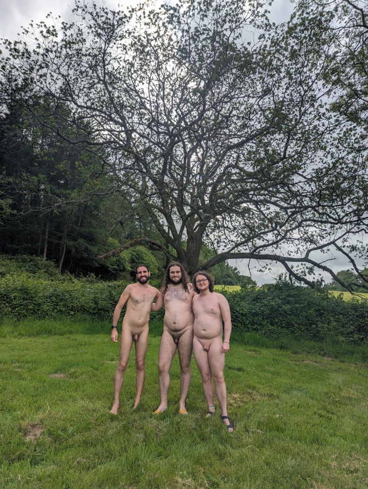 From left to right, Vinc, Ivan (me) and David from the Spanish Naturist Federation are standing naked and smiling at the camera. Vinc is a member of ANVA (Asociación Naturista Valenciana), while David and Ivan are members of CCN (Club Català de Naturisme). In the background there are green meadows and forests, and a large branchy tree occupies a central place.