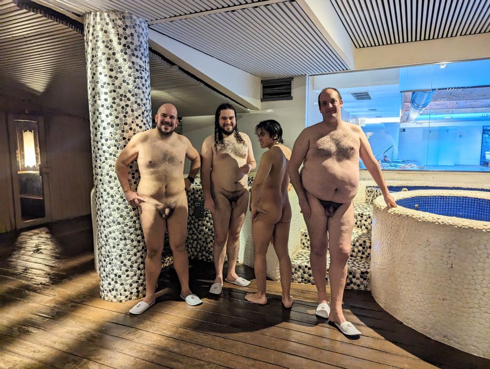 Me, second from the left, standing nude with David, Guillem, and Marc from the Young Naturists group in a spa in Barcelona. A small round pool with a white mosaic edge is beside us, and warm lighting reflects off the pebble-covered pillars and the wooden floor.