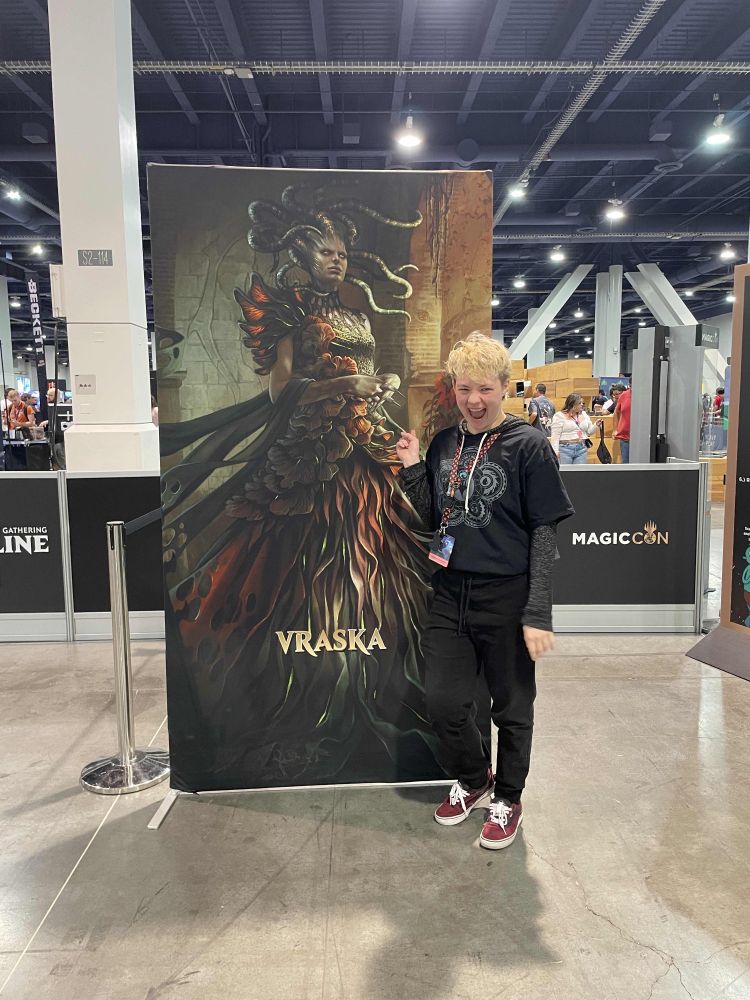 picture of me standing next to a 7 foot tall poster of Vraska. I am pointing at her and smiling really big.