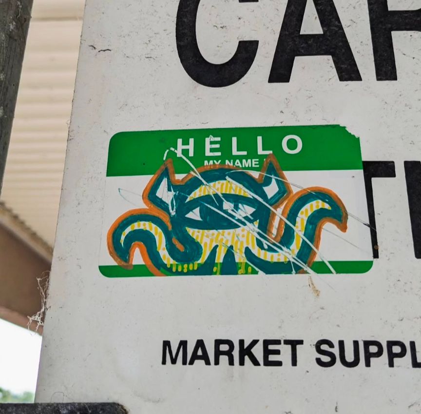 A green Hello My Name Is sticker stuck to a cart corral sign with a drawn yellow Vacant covering the white write-in space.