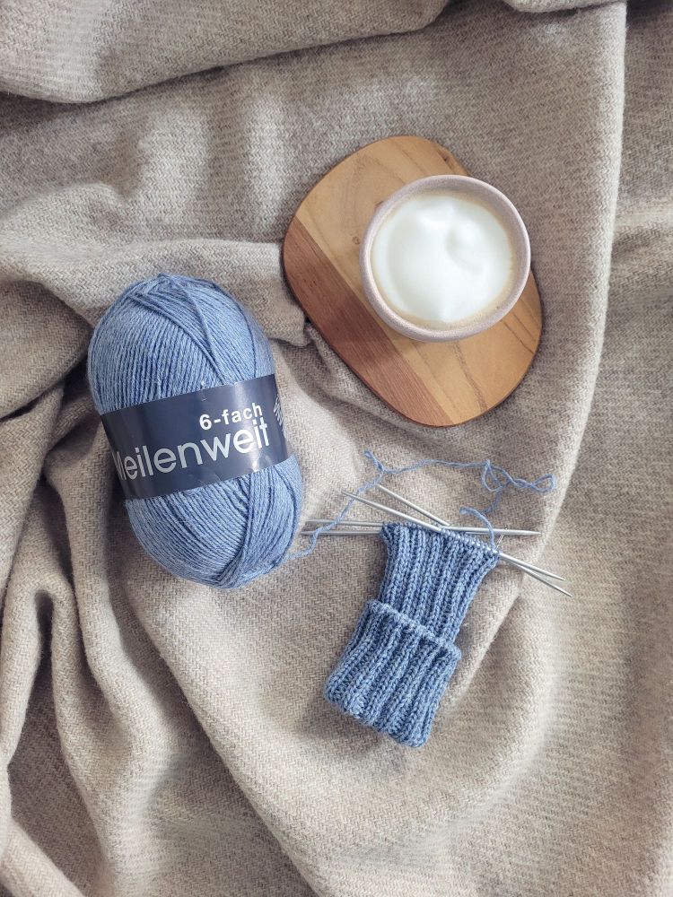 Displayed on a brown woolen blanket are a ball of jeans blue DK weight sock yarn attached to half knitted sock and a cappucino. 