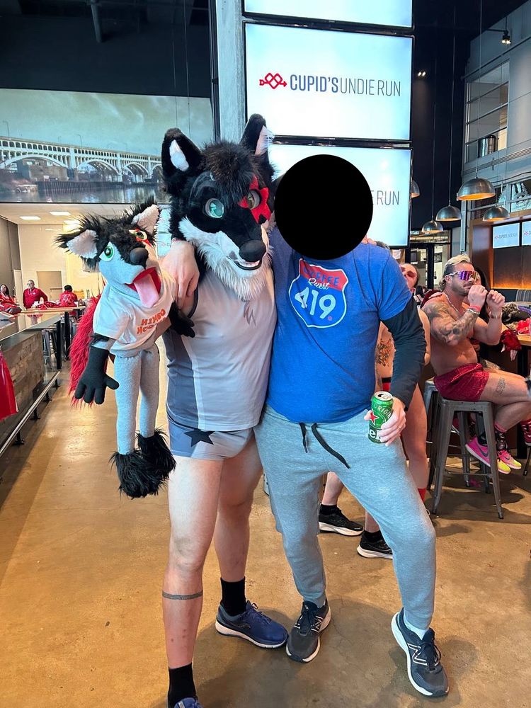 Forrest Husky's current fursuit is black with grey markings beneath the muzzle. There is a red star marking surrounding his left (not the viewer's left) eye. In public, he often carries around a "mini-me" puppet named Lil Forrest that looks like his fursuit. The puppet has been used in sexual acts. I have covered the face of the person posing beside him for privacy.