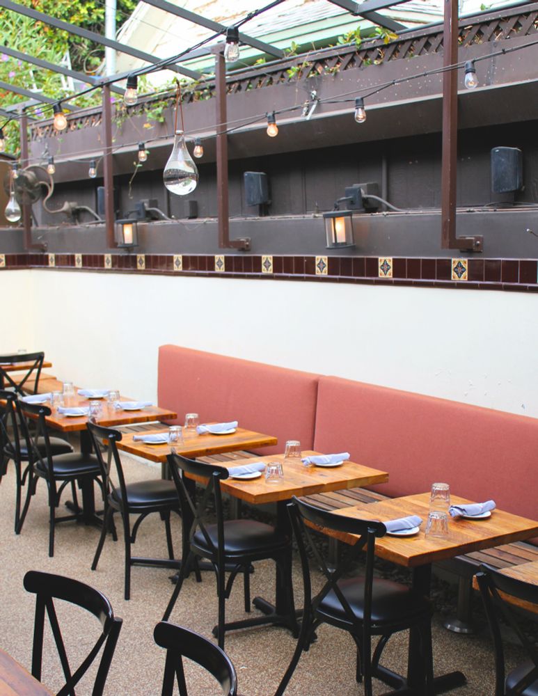 An airy outdoor patio on a bright afternoon. The patio has white walls with brown tiled accents, warm sconce lights, suspended bulb lights, rows of wood tables with black chairs, and an inviting communal wooden bench with a plush orange back rest on one side. Each table is set with water glasses turned upside down and small ceramic plates with silverware encased in linen napkin “roll-ups” on top.