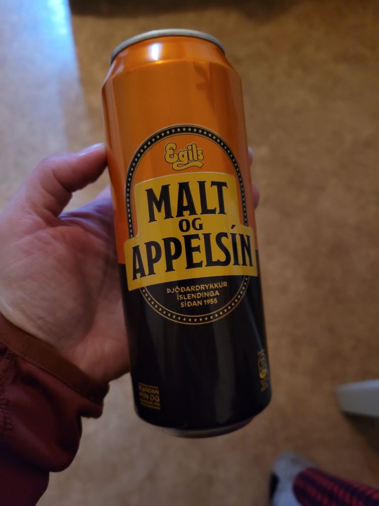Jemand hält eine Getränkedose in der Hand, halb orange, halb braun. Die Aufschrift sagt "MALT OG APPELSÍN"