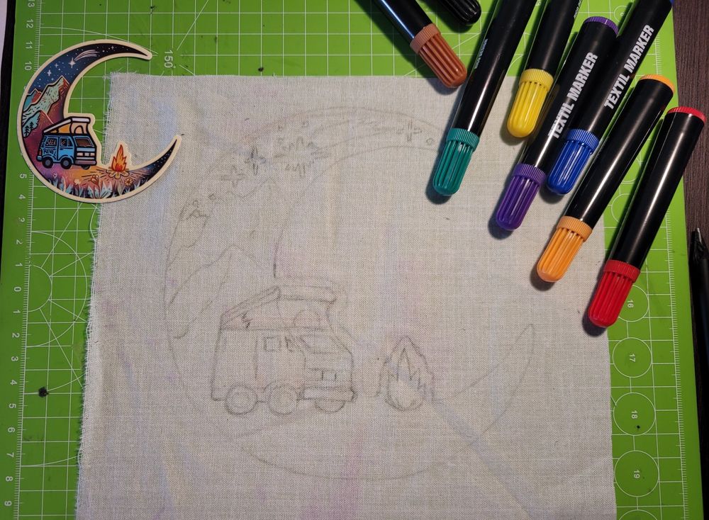 Ein Halbmond-Sticker mit Van und Lagerfeuer in den Bergen liegt auf einem weißen Stoffstück, auf das das Motiv mit Bleistift aufgezeichnet ist. Am oberen Rand liegen Textil Marker