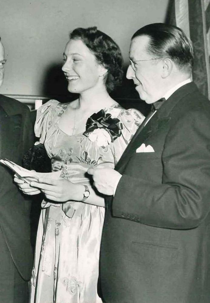 Caroline Herzenberg, age 17, as a finalist in the 1949 Science Talent Search contest. The photo is black and white. She is wearing a long dress or gown, light colored, with puffy sleeves and a bow pinned over her heart. She has short, dark hair. Her head is turned to the side and she is smiling at someone off camera.