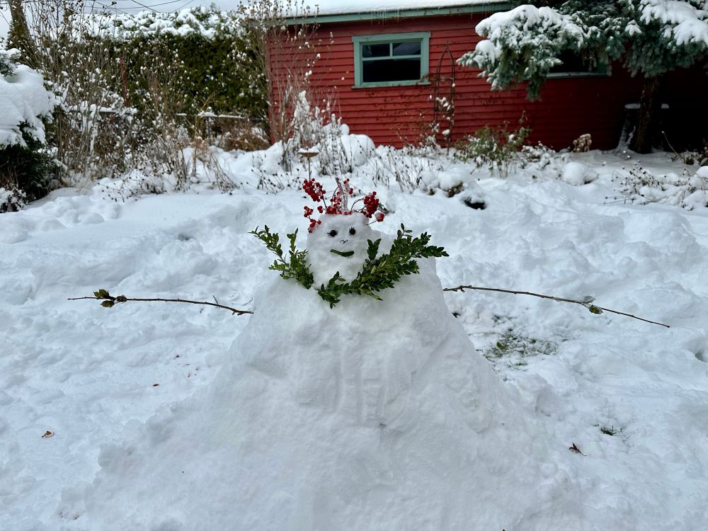 A lumpy snow woman with a laurel wreath and a crown of bright hawthorn berries. She has a leaf smile and two dried flowers for eyes. The overall shape is more mound than cone, certainly not the traditional stacked snowball form.