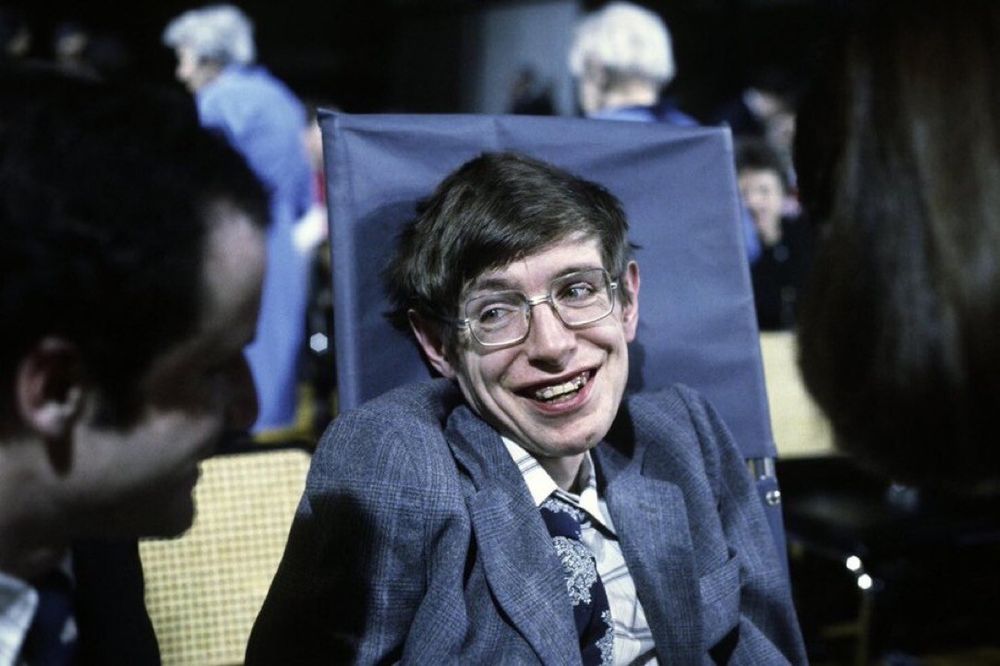 Color photo of Stephen Hawking. He is seated in his wheelchair, smiling at someone to his right. Hawking has dark hair and is wearing glasses. He has on a dark blue jacket, light plaid shirt, and a patterned blue tie.