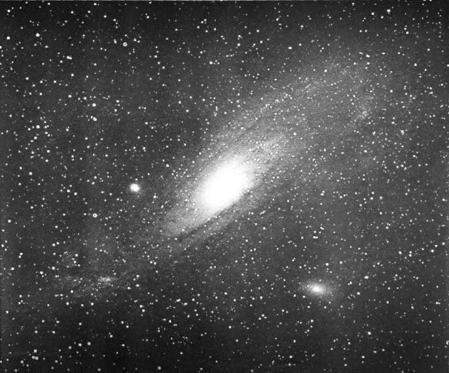 Black and white photo of the Andromeda Galaxy, a spiral galaxy with a strong central bulge. Two smaller satellite galaxies float nearby, and the foreground is dotted with stars.