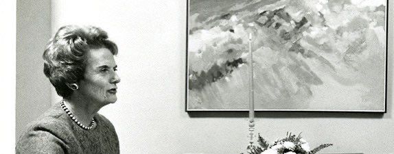 Black and white photo of Mina Rees. She is older, possibly in her early 60s, wearing what appears to be a gray wool top and a string of pearls. Her hair is short, and she is in front of a wall with a large abstract painting. She is seen in profile, looking to the right of the photo.