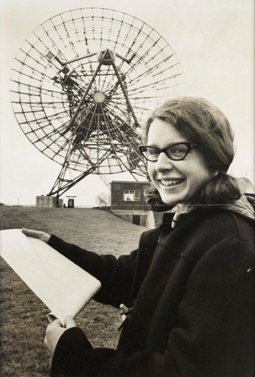 A black and white photo of Jocelyn Bell Burnell looking at a printout in front of the large radio dish she used. She is wearing cat's-eye glasses with her brown hair pulled back. Her head is turned towards the camera and she is smiling.