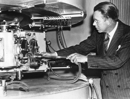 A black and white photo of Ernest Lawrence working on part of his cyclotron. Despite working in the lab he is wearing a pinstripe suit with a double-breasted jacket -- a very formal affair by today's standards, totally lab-to-evening wear. There is a white handkerchief in the breast pocket of his jacket. Lawrence his looking away from the camera, at the apparatus that he is working on. He is wearing glasses and his hair is slicked back. The apparatus appears to be two large cylinders (probably supporting magnets) with machinery situated between them.