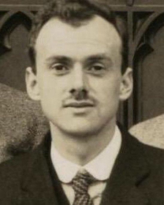 A black and white photo of Paul Dirac in his early 20s. He is looking directly at the camera, and while not smiling he does look pleased. Dirac is wearing a dark jacket and white collared shirt. His tie sports a stairstep strip pattern. Dirac has a thin mustache.