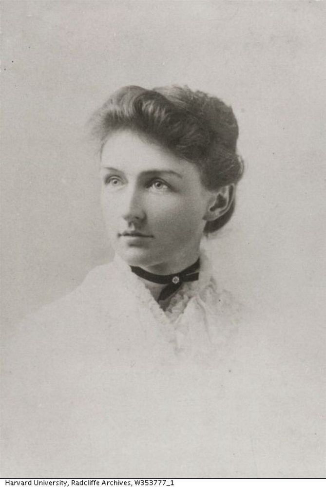 Black and white photo portrait of Annie Jump Cannon. She is wearing a white blouse or shirt with a frilly collar that fades into the white background of the photo, and a dark choker around her neck. Her hair is pulled back and she is looking to the left of the camera.