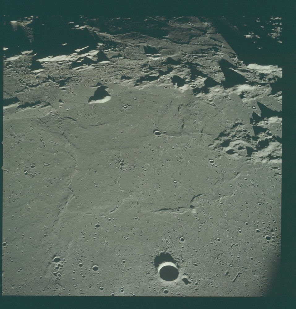 Color photo of the lunar surface. It is a gray-tan color. This square patch has a perfectly round little crater near the bottom of the photo, along with what appears to be rocky, mountainous terrain near the top. The inside of the crater is in shadow; the hills near the top cast long shadows on the terrain behind them.