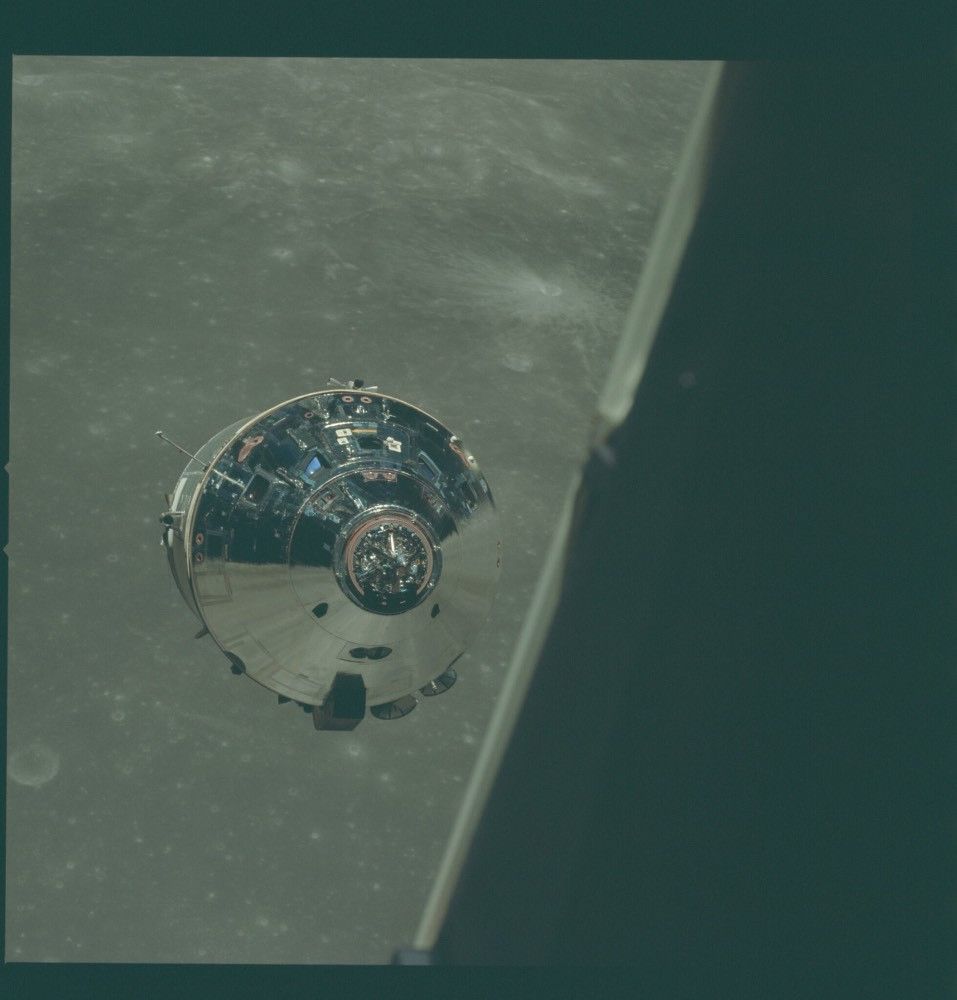 The Command and Service Module, as seen from the Lunar Module. It is cylindrical, with a conical top that is highly reflective and shows an image of the surface below.