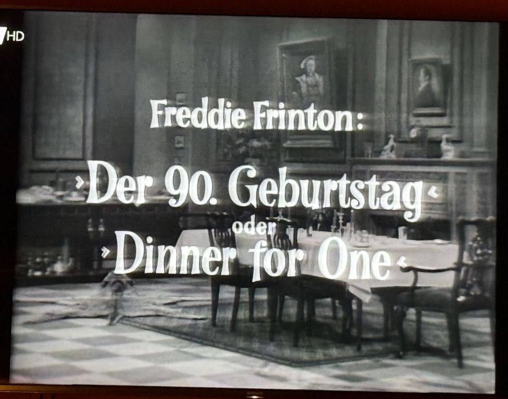 A black and white title card over an elaborate dinner table set for five, reading „Freddie Frinton: Der 90. Geburtstag oder Dinner For One“