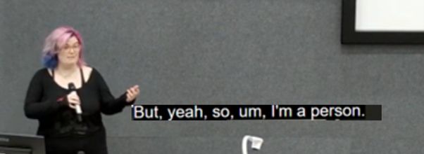 Image of Rosie (pink and blue shoulder-length hair, pale skin, plus-sized, in a long black top with the D&D logo), holding a microphone in one hand and gesturing with the other. Subtitles on the image from audio transcription of Rosie's lecture say 'But, yeah, so, um, I'm a person.'
