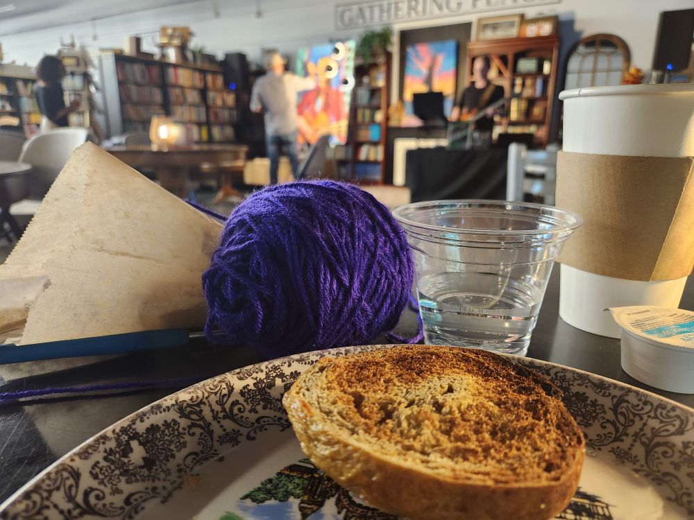 View from a table with a pumpkin bagel, coffee cup, and yarn in the foreground while an artist paints and a musician performs in the background at a community event.