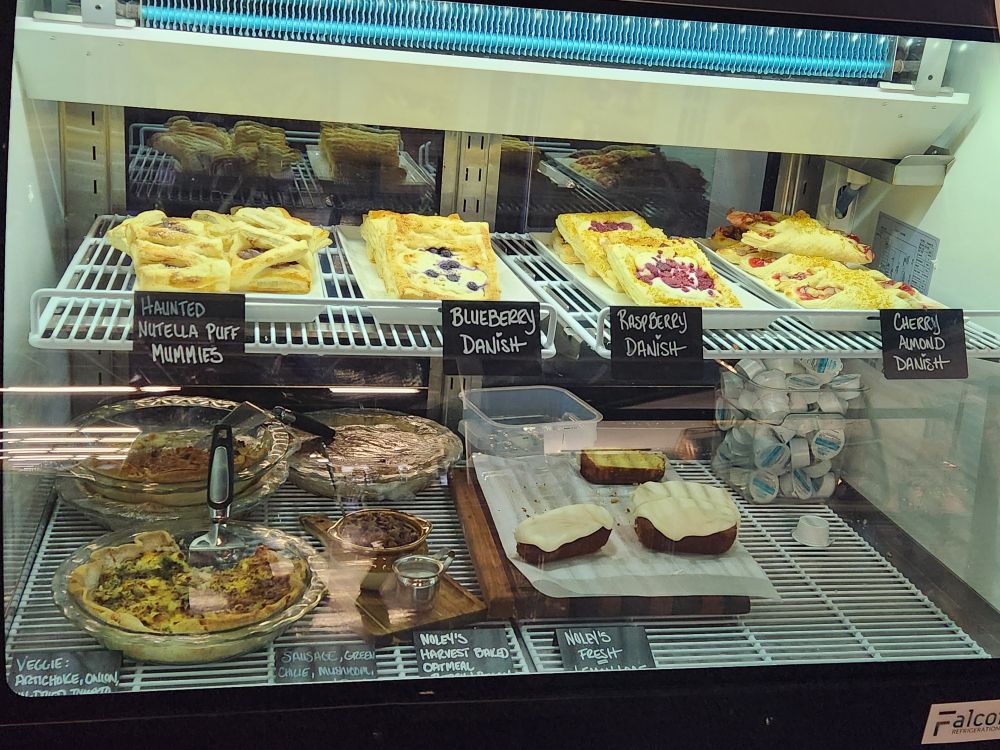 Display case with pastries including Nutella puff pastries, blueberry Danish, raspberry Danish, and cherry almond Danish labeled on trays inside a refrigerated case.
