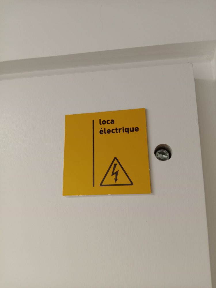 L'armariet del quadre elèctric té un cartell quadrat groc amb un triangle i un llamp (perill de descàrrega elèctrica) i el text 'loca électrique' a sobre.