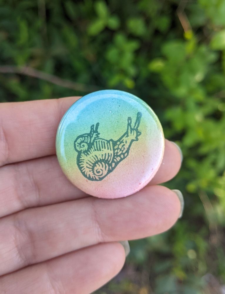 A hand holding a handprinted 1.25" pinback button, printed with a rainbow roll and a tiny snail-on-snail hand carved stamp