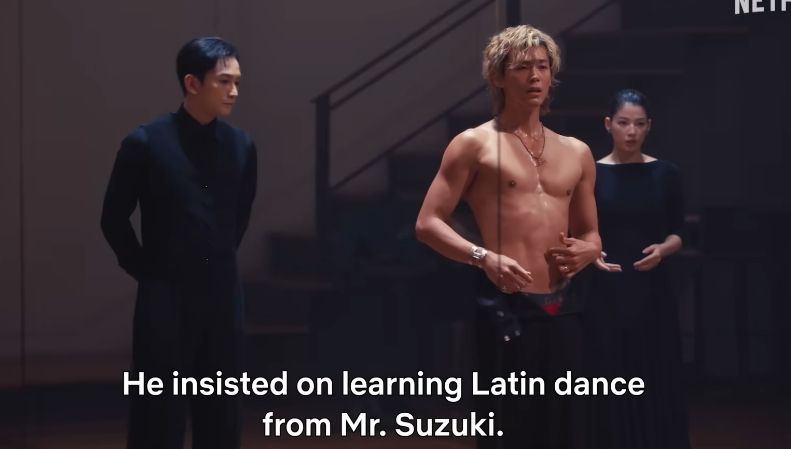 homem de terno preto olhando pro lado, o personagem sem camisa com a cueca meio de fora da calça aberta, atrás uma moça de preto. legenda he insisted on learning latin dance from mr suzuki