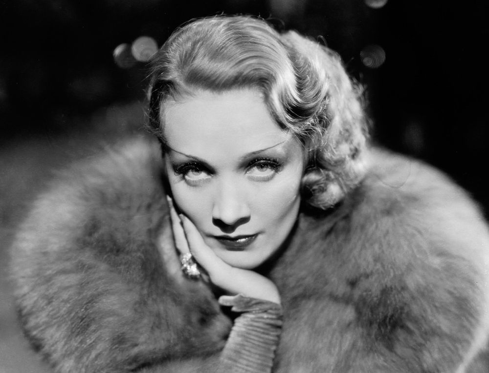 Blackand white photo of actress Marlene Dietrich with her chin resting on her hand. She is staring confidently and questioningly into the camera. She is wearing soft furs. Only her head and shoulders are shown against a black background.