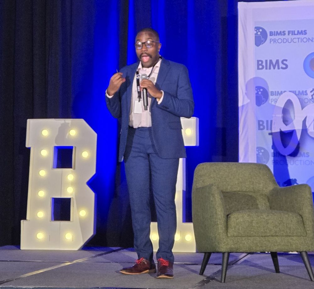 A Black man is on stage in a dark blue suit with a light pink shirt and brown shoes with red laces. He is holding a microphone in his left hand. 