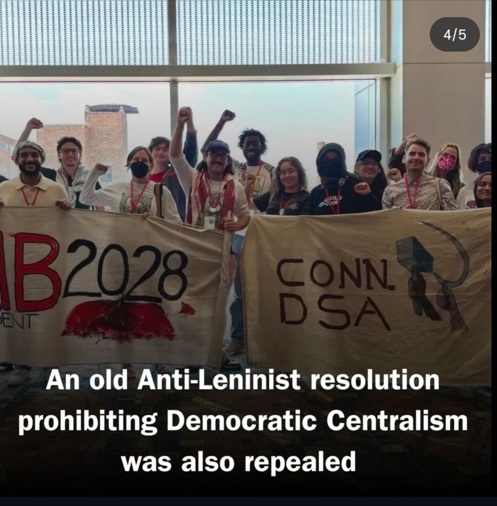 DSA members posing for a photo, some with raised fists, some are wearing masks that appear to be cloth. The ones in the front of the group are holding banners ike reads 2028 and the other reads Conn DSA (convention DSA) with two fists. One holding a hammer and the other holding a sickle. Notably in the background there are two large windows providing natural light and a view of some of Chicago's skyline. 