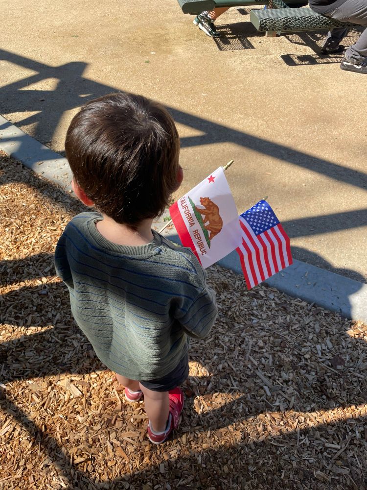 A dark haired toddler in a green shirt holding small California and US flags